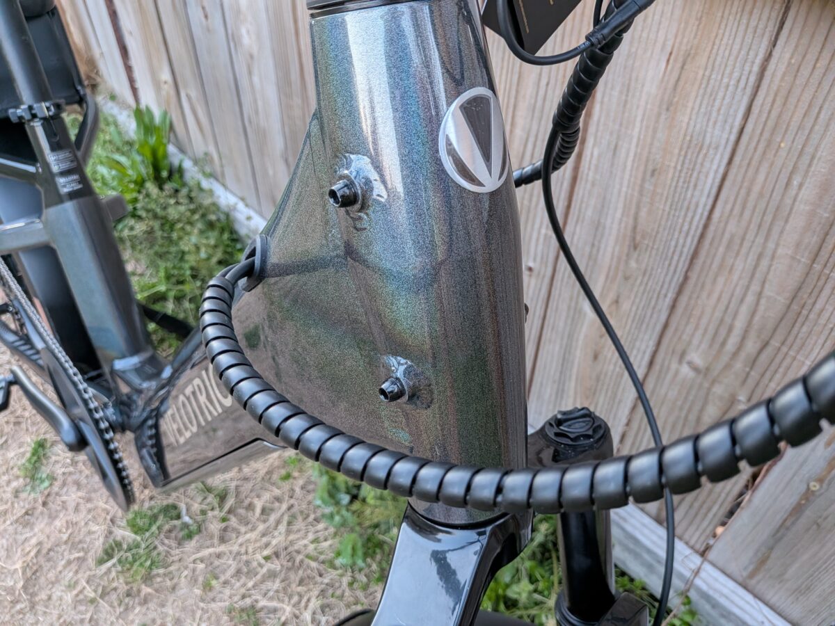 Close-up of the Velotric GoMad head tube with a metallic gray frame, visible cables, and headset area at the front of the bike near a wooden fence.